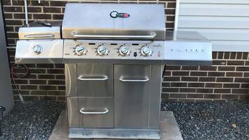 A stainless steel outdoor grill with four burners and a side burner, mounted on a brick wall with a utility box nearby.