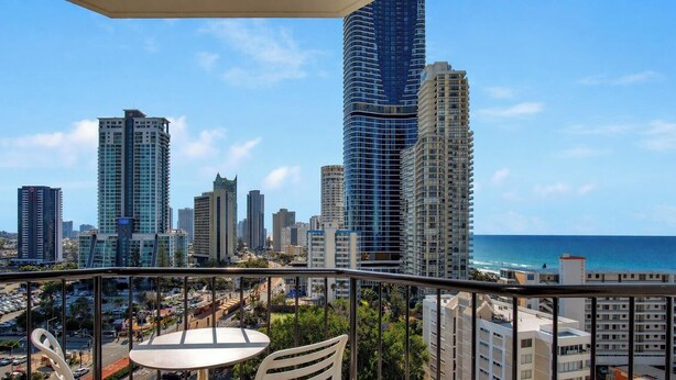 A balcony with a table and chairs, overlooking a cityscape with tall buildings and a view of the ocean.