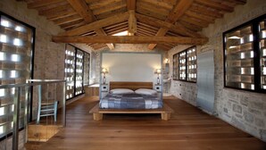 A spacious bedroom with a wooden ceiling, stone walls, a large bed, and a view through a glass door. - Villa Fontanine -  Poggibonsi (SI)