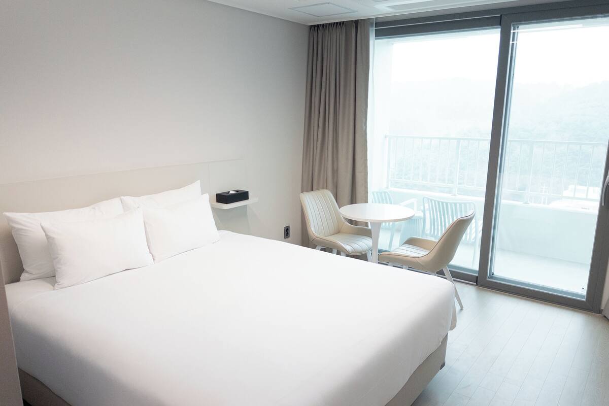 A hotel room with a large bed, a small round table, a chair, and a balcony with a view.