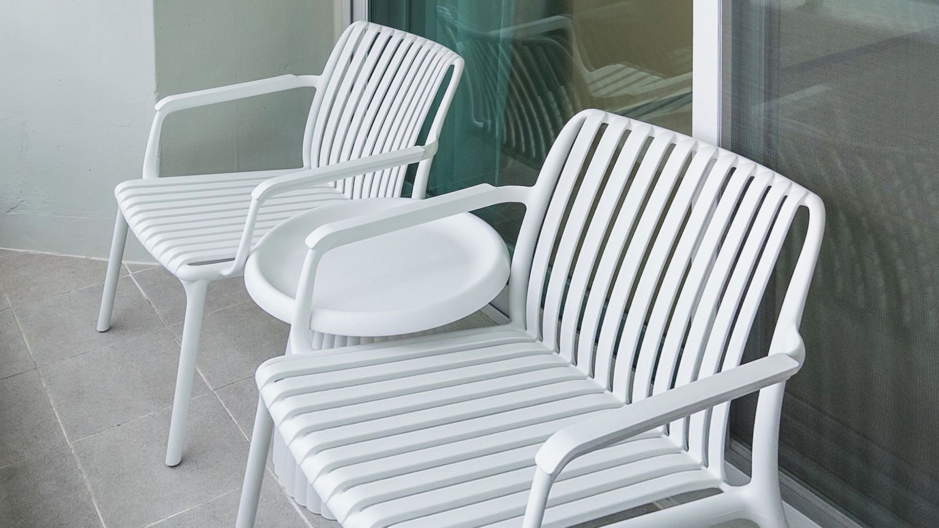 Three white plastic chairs with a ribbed backrest and armrests, positioned on a tiled floor.