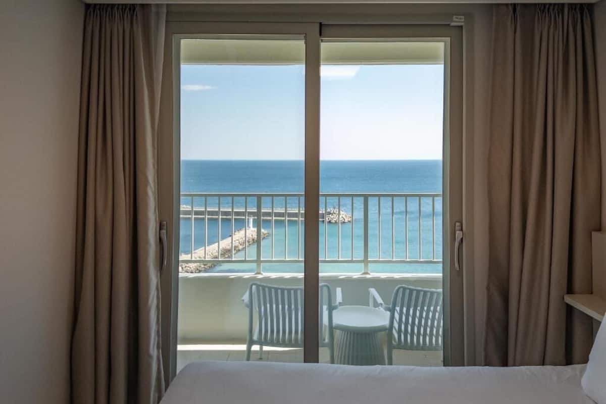A hotel room with a balcony overlooking the sea, featuring a bed, two chairs, and a small table.