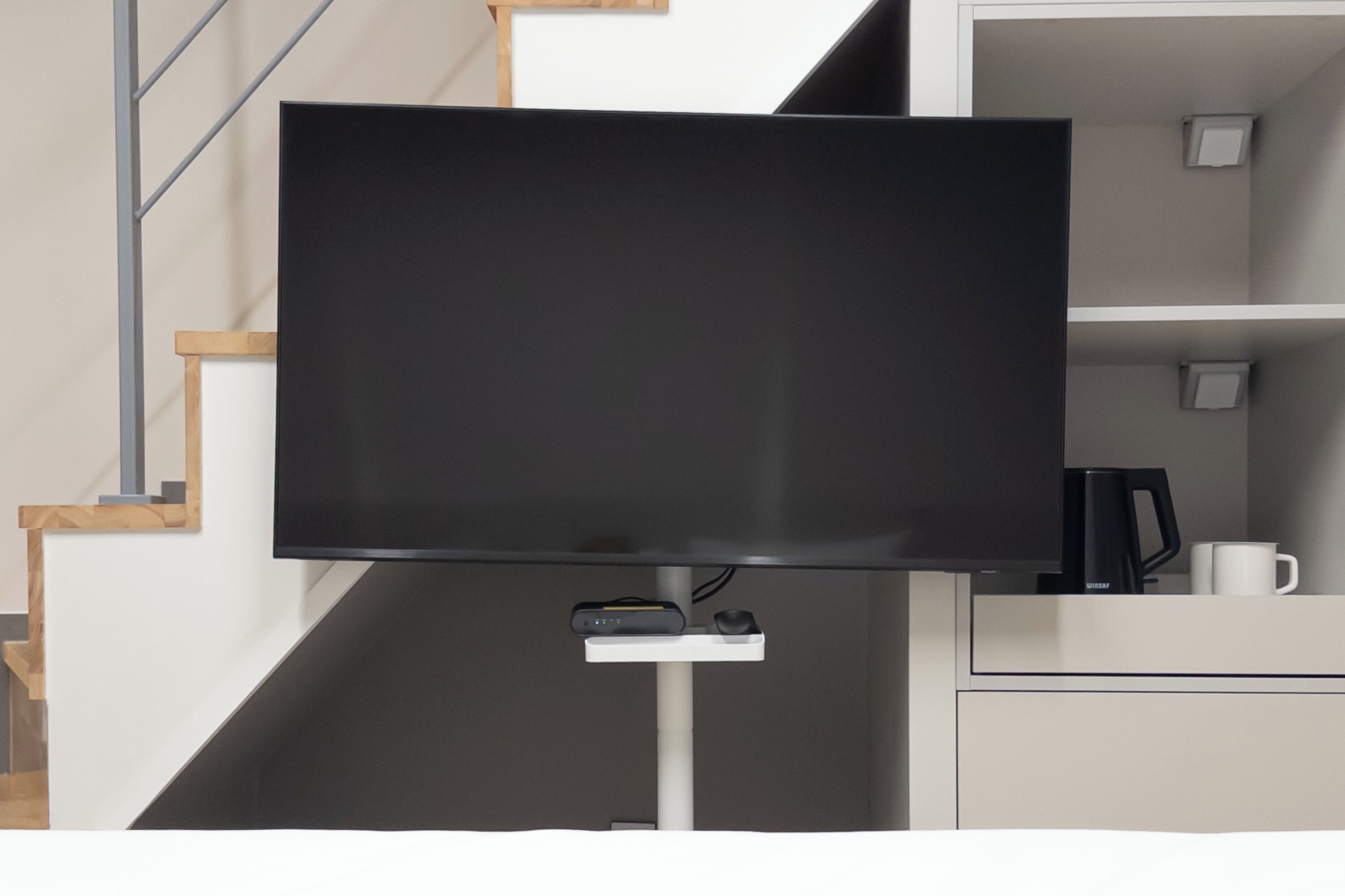 Un televisor de pantalla plana montado sobre un soporte en una habitación moderna con una escalera y un mueble blanco con cafetera.