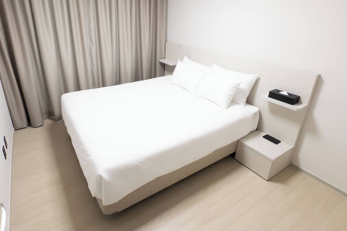 A neatly made bed with white linens, a bedside table with a remote control, and a small storage box.