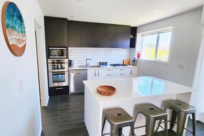A modern kitchen with dark cabinets, white countertops, stainless steel appliances, and a large island with stools. - Spacious 5BR 2BA plus sleepout & games room Family Home (Christchurch)