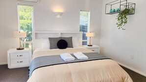 A neatly arranged hotel room with a bed, bedside lamps, a nightstand, a wall-mounted shelf with plants, and a window with blinds. - Spacious 5BR 2BA plus sleepout & games room Family Home (Christchurch)