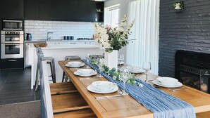 A modern kitchen with dark cabinetry, white tiles, and a long wooden dining table set for six with plates, glasses, and a vase of flowers. - Spacious 5BR 2BA plus sleepout & games room Family Home (Christchurch)