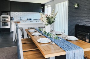 A modern kitchen with dark cabinetry, white tiles, and a long wooden dining table set for six with plates, glasses, and a vase of flowers. - Spacious 5BR 2BA plus sleepout & games room Family Home (Christchurch)