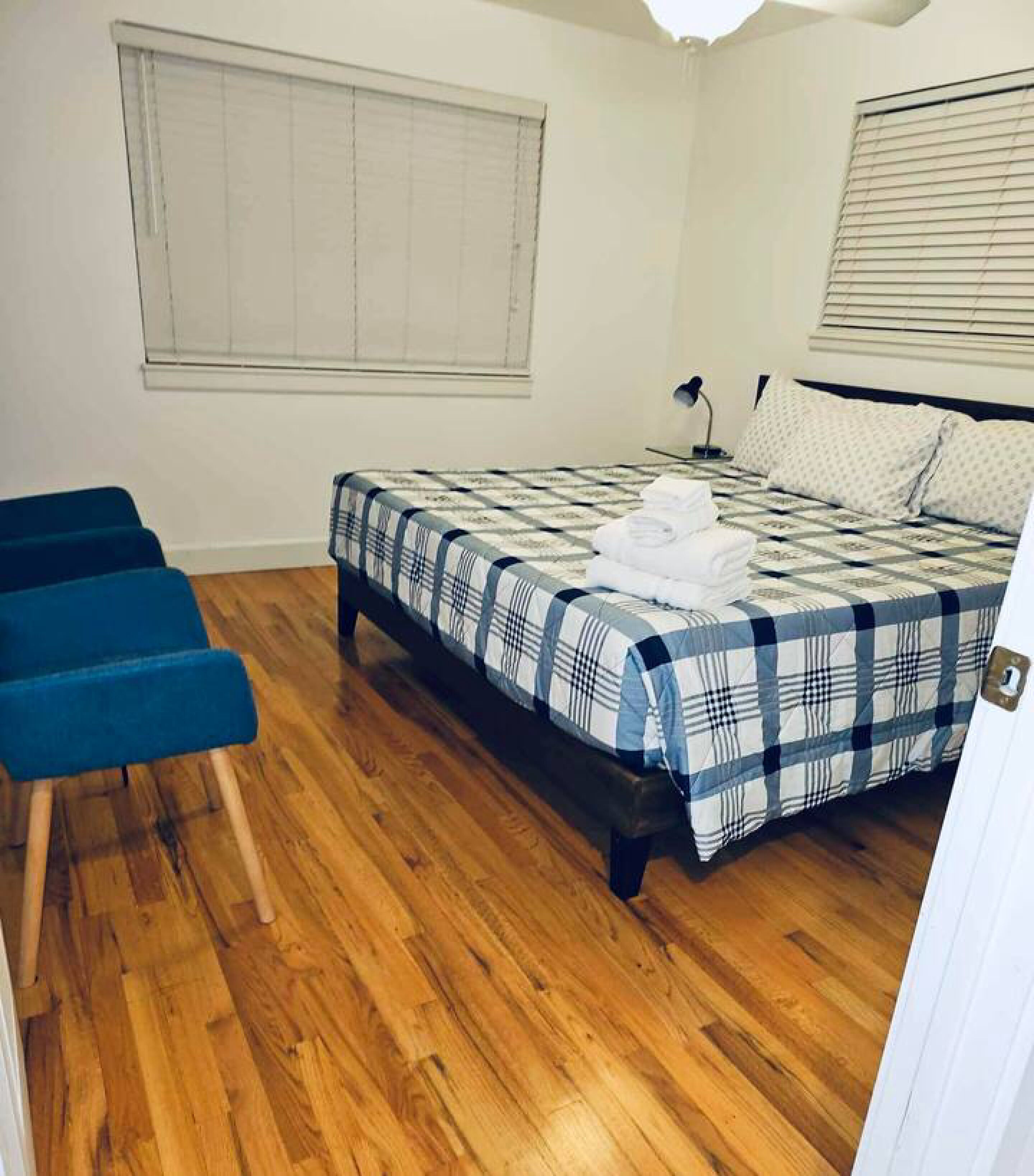 A bedroom with a bed, a blue chair, wooden flooring, and a window with blinds.