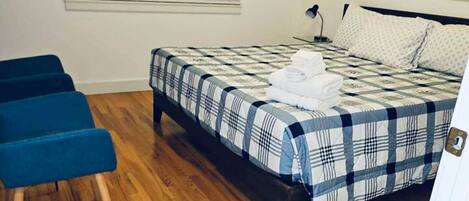 A bedroom with a bed, a blue chair, wooden flooring, and a window with blinds.