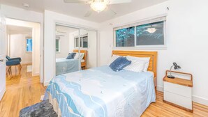 A bedroom with a bed, a nightstand, a ceiling fan, a window with a view, and a door leading to another room.