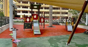 Un parque infantil con toboganes y estructuras para escalar, rodeado por una zona vallada y un edificio al fondo.