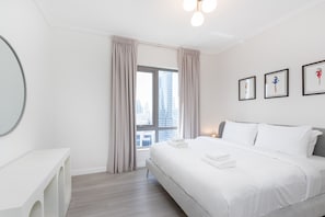 A modern hotel room with a large bed, a bedside table, a window with curtains, and framed artwork on the wall.