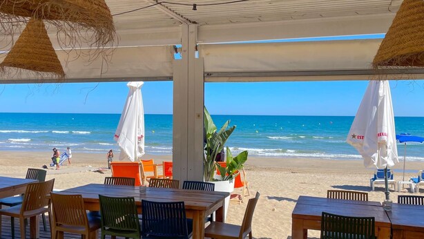 An outdoor seating area with wooden tables and chairs, white umbrellas, and a view of the beach and ocean.