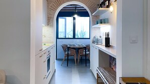 Uma cozinha estreita com um arco que dá para uma área de refeições. Há vigas de madeira no teto, uma janela com vista e uma prateleira com utensílios de cozinha.