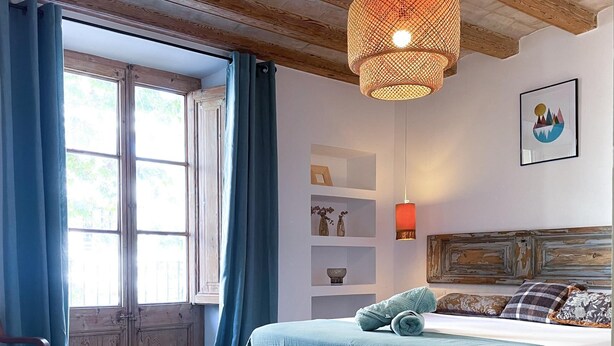 Quarto com cabeceira de madeira, cama com colcha azul-turquesa, uma cadeira azul, guarda-roupa de madeira e uma janela com cortinas azuis.
