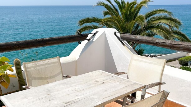 A balcony with a table and chairs overlooking the sea, palm trees, and a wooden railing.