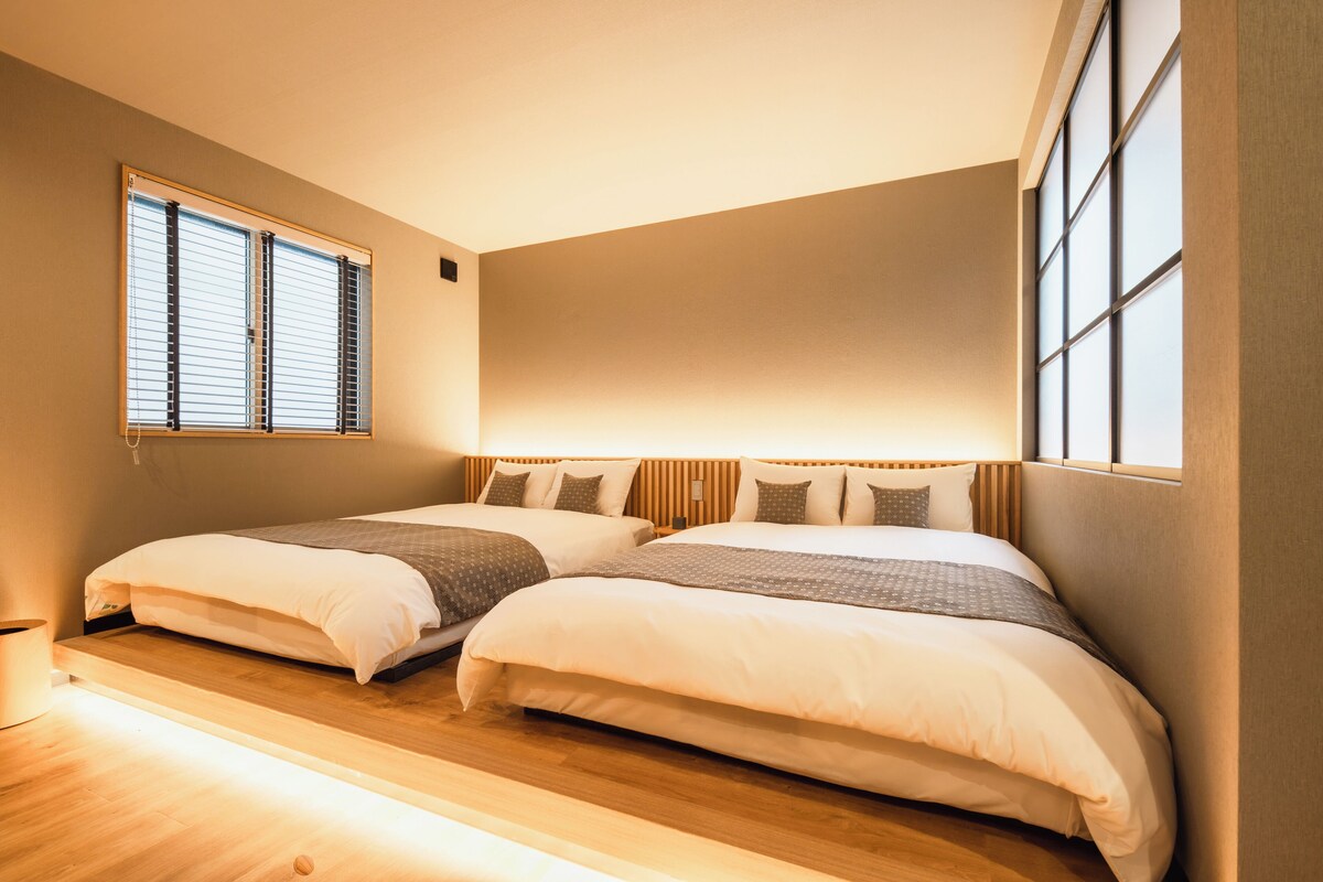A bedroom with two beds, wooden flooring, and large windows with blinds.