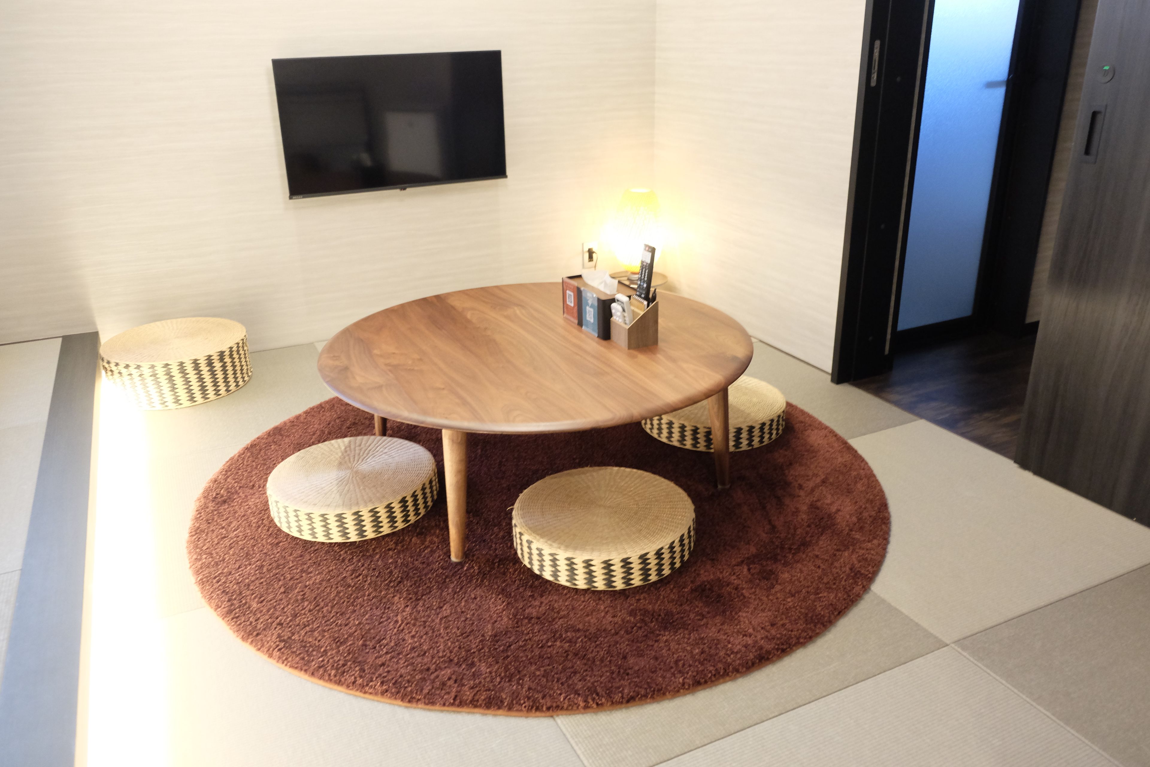 A round wooden table with a woven rug, four matching stools, a wall-mounted TV, and a small table with various items.