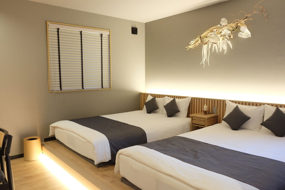 A hotel room with two beds, a bedside table with a lamp, a wall-mounted heater, and a window with blinds.