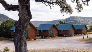 A row of wooden cabins with green doors and blue roofs, set against a backdrop of hills and trees.