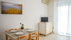 Un coin repas avec une table dressée pour quatre personnes, un téléviseur posé sur un meuble et un tableau représentant une scène de plage accroché au mur.