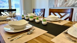 Una mesa de comedor dispuesta con cuencos, platos y cubiertos blancos, rodeada de pequeñas macetas con plantas.