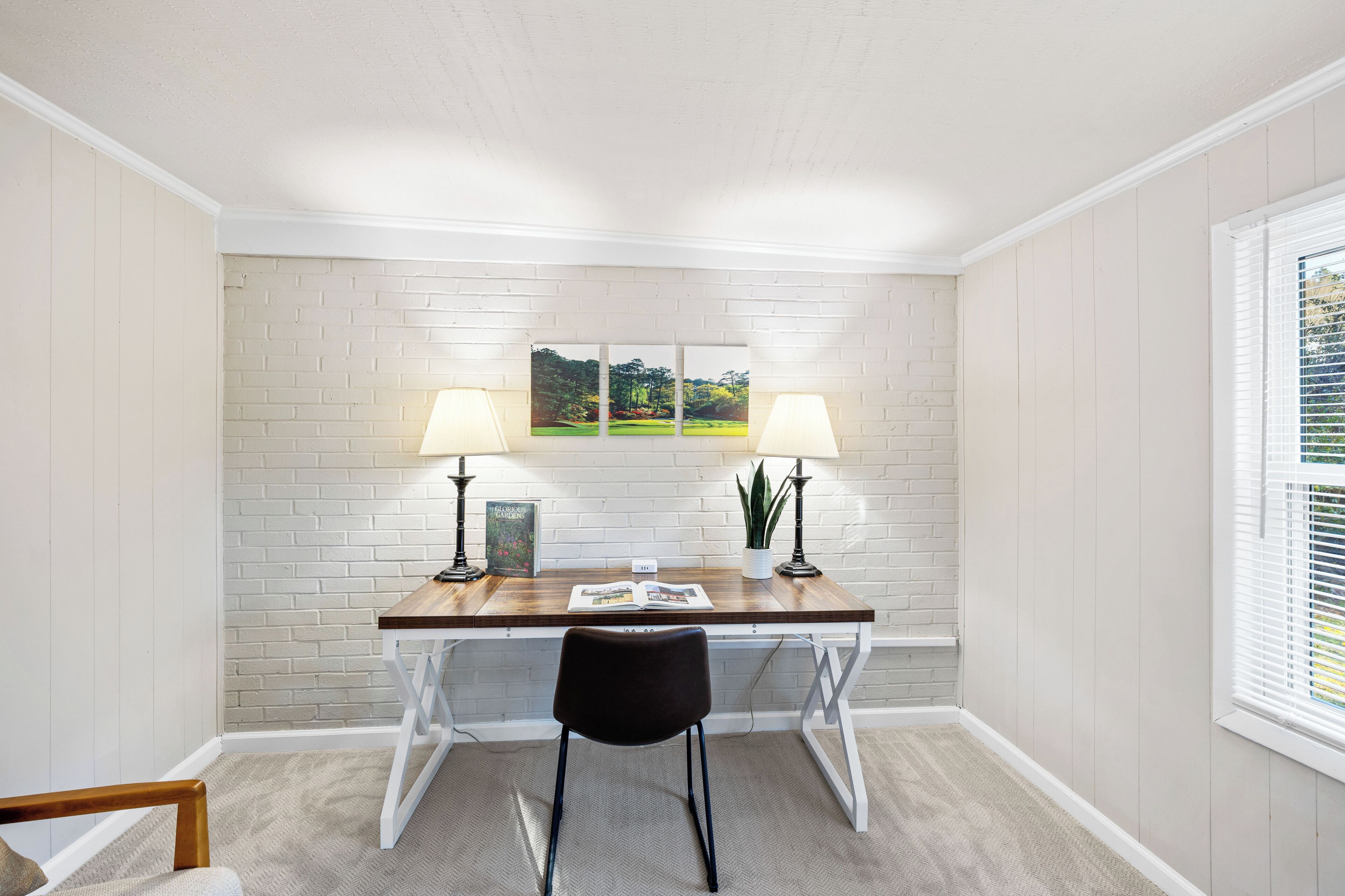 Une pièce avec un mur en briques blanches, un bureau en bois avec deux lampes, une chaise et une fenêtre avec des stores.