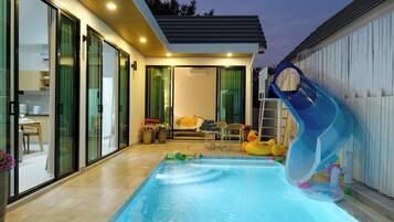 A swimming pool with a slide, a dining area with chairs and a table, and a bedroom with a bed and curtains.
