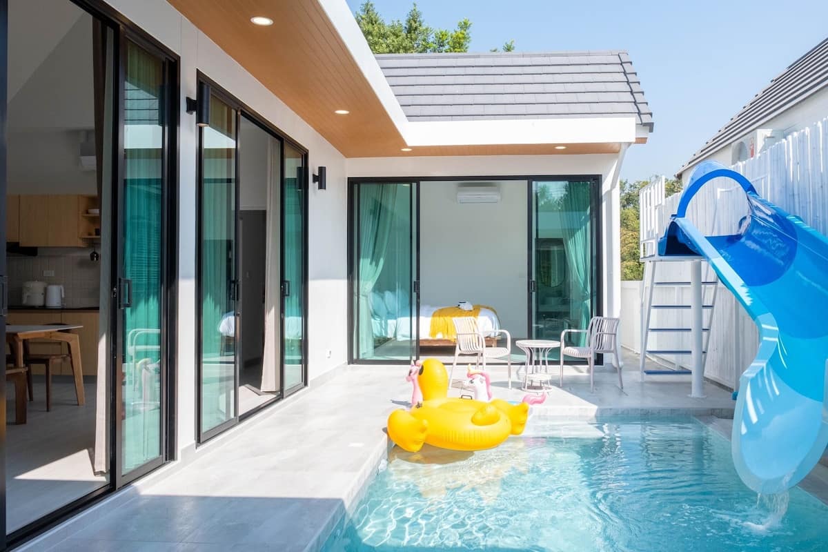 A modern outdoor pool area with a slide, a yellow inflatable pool float, and a bed visible through the glass doors.
