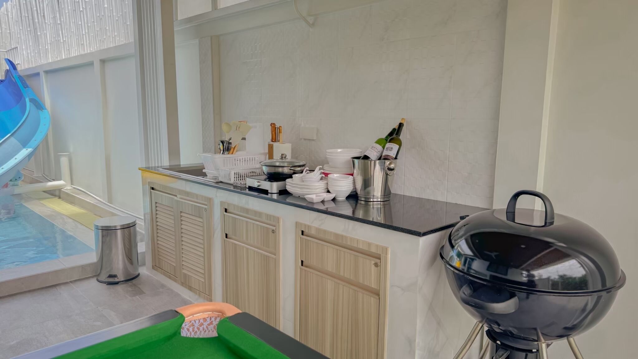 A modern kitchen with a poolside area, including a barbecue grill, a sink, and a variety of kitchenware.