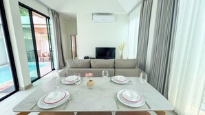 A dining area with a table set for two, a sofa, a TV, and a view of a pool. - Wana Villa ChaAm (Cha-am)