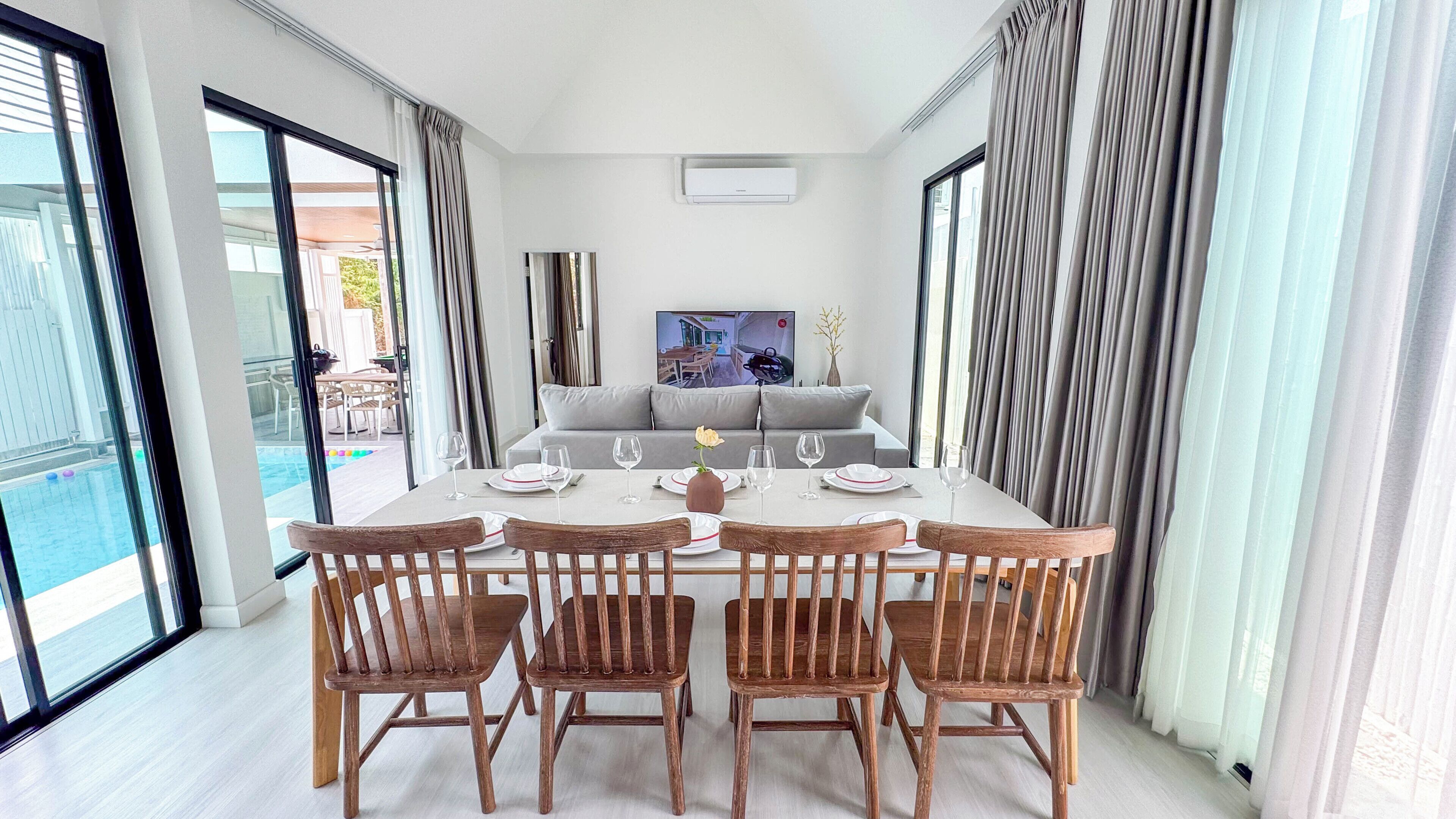 A dining area with a table set for six, six wooden chairs, a grey sofa, and a large window with curtains.