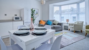 A modern living room with a dining area, featuring a white table set for four, a grey sofa with colorful cushions, a large window with blinds, and a white cabinet with a microwave.
