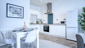 A modern kitchen with white cabinets, a stainless steel range hood, a microwave, and a refrigerator. There is a dining table with chairs and a framed picture on the wall.