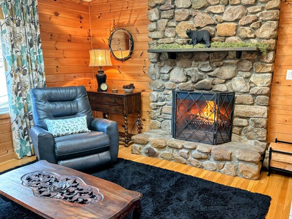 A cozy room with a stone fireplace, a leather armchair, a wooden coffee table, and a floral curtain.