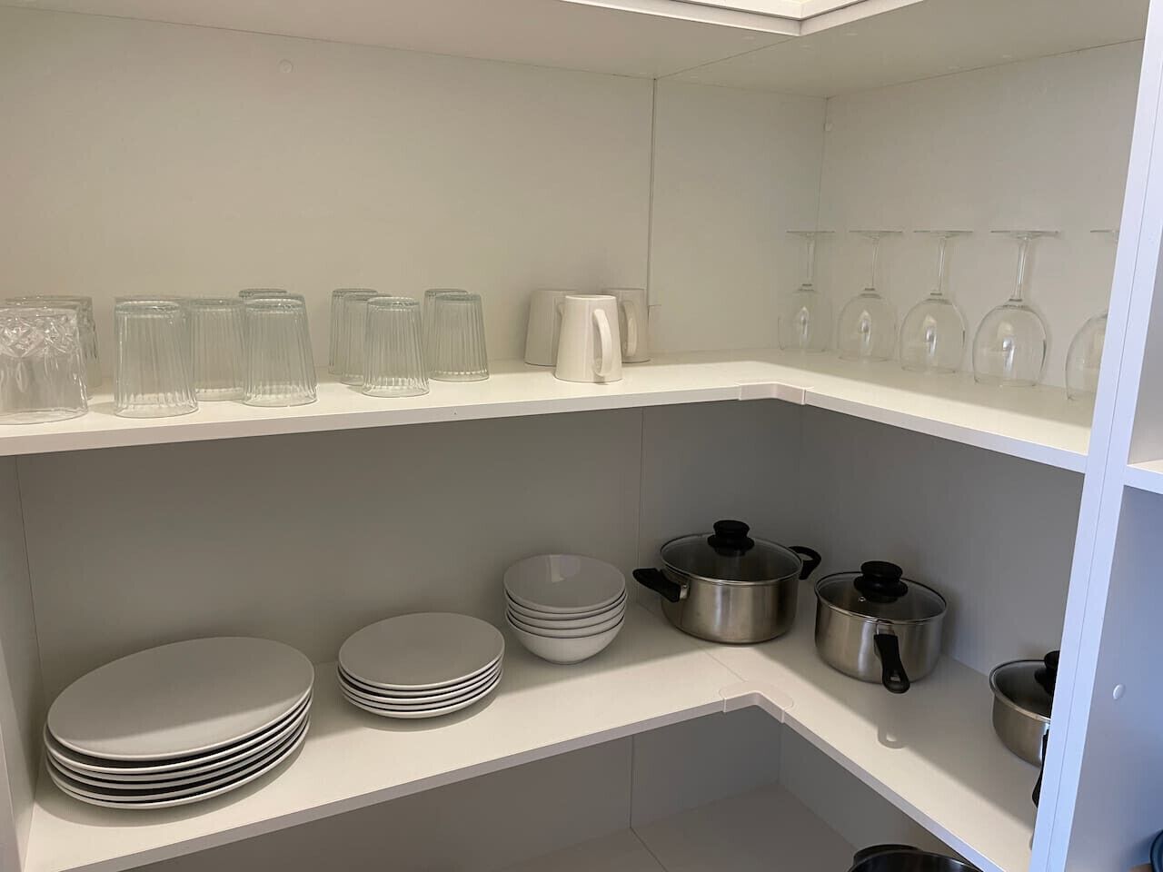 Una cocina con estantes blancos, platos, tazones y ollas de acero inoxidable apilados.