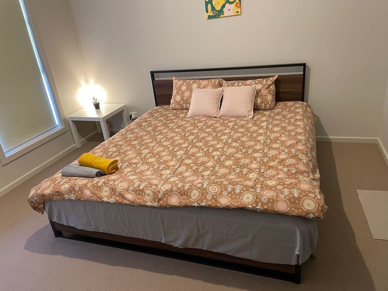 Een netjes opgemaakt bed met beddengoed met een patroon, een nachtkastje met een lamp en een raam met jaloezieën.