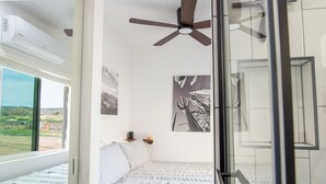 A bedroom with a bed, a ceiling fan, a window with a view of the outdoors, and a wall-mounted artwork.