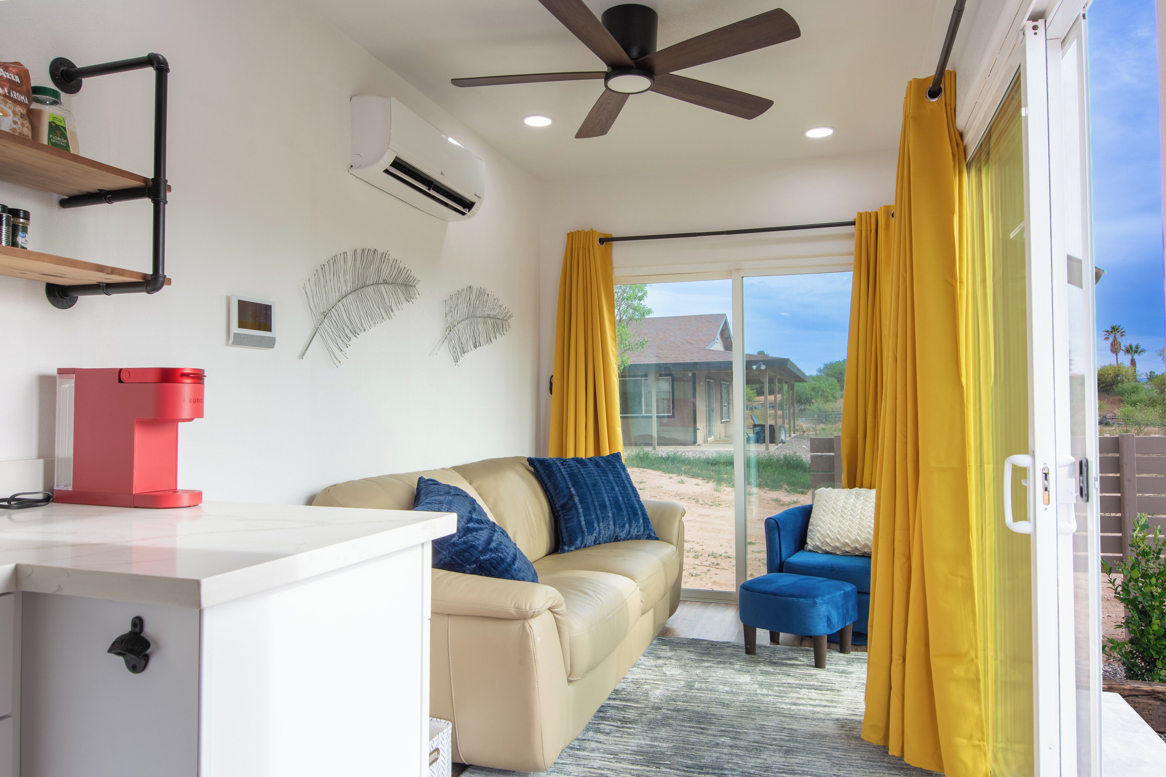 Una sala de estar moderna con un sofá color beige, una cafetera roja, un ventilador de techo y un amplio ventanal corredizo con cortinas amarillas.