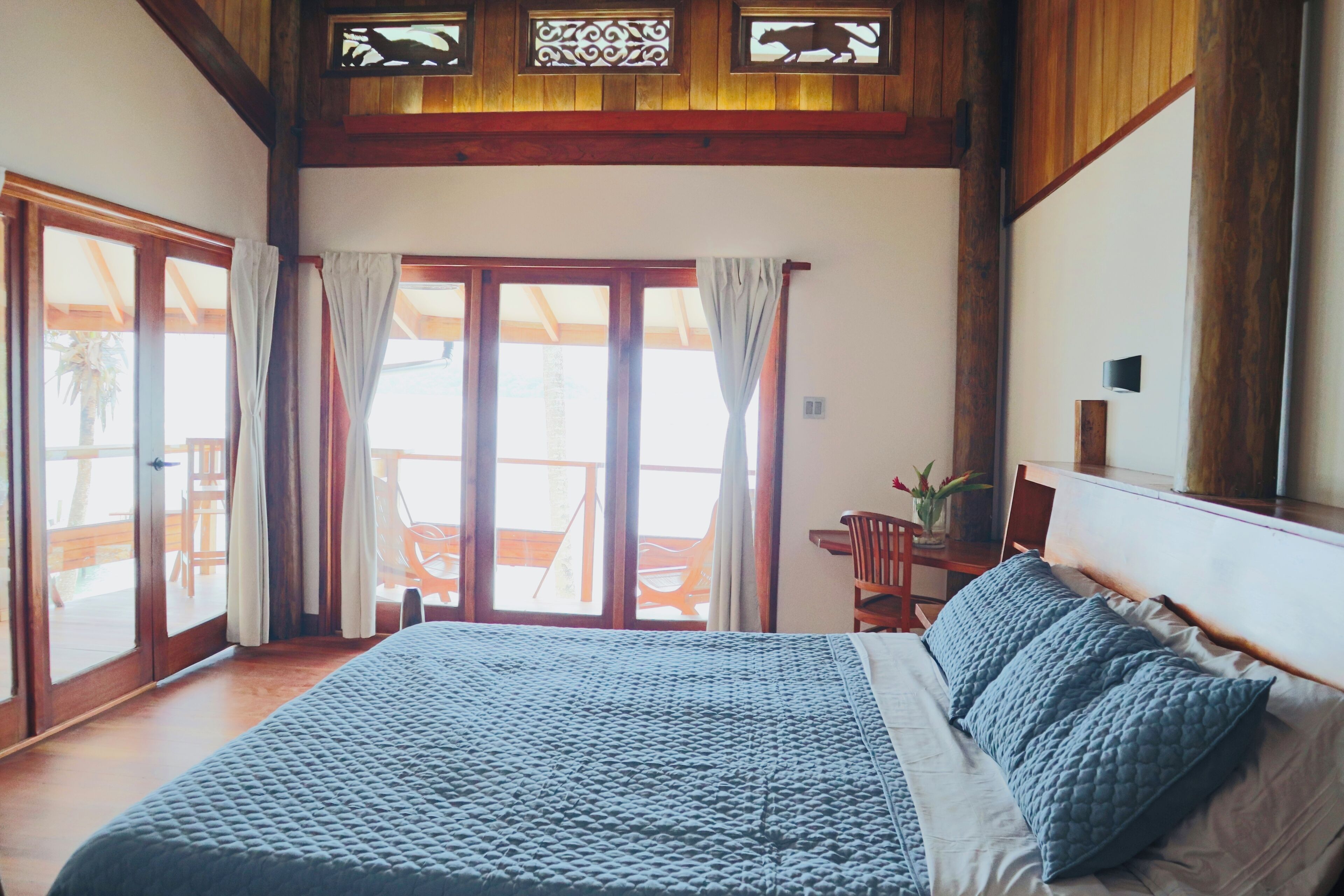 Un dormitorio con una cama grande, vigas de madera en el techo, un ventanal corredero que da a un espacio exterior y una pequeña mesa con sillas.
