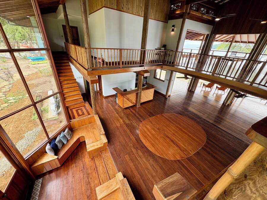 Un interior espacioso con suelo de madera, una zona de estar central y una escalera que conduce a un nivel superior.