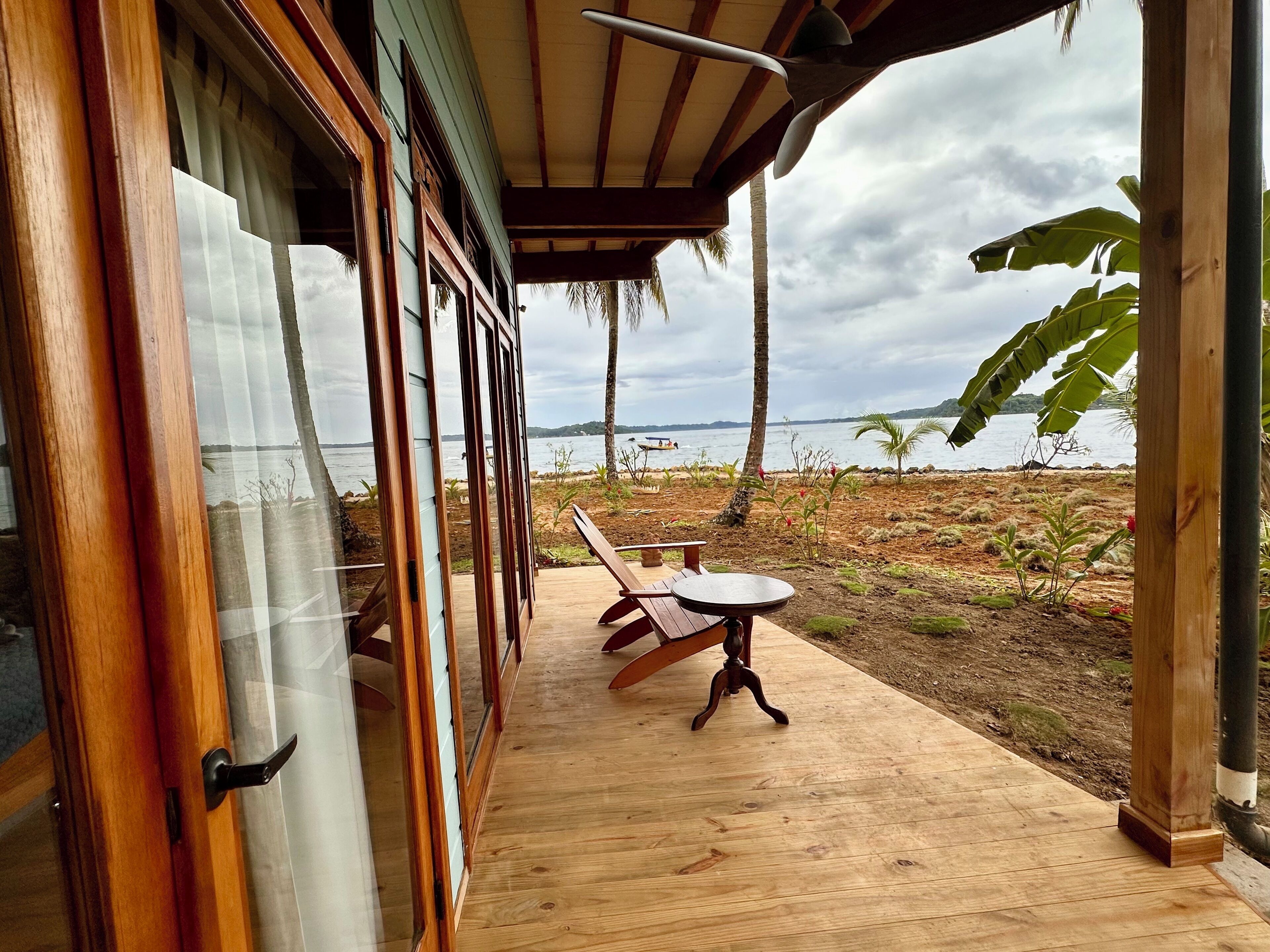Une terrasse en bois avec une table et des chaises, donnant sur une plage bordée de palmiers et offrant une vue sur la mer.