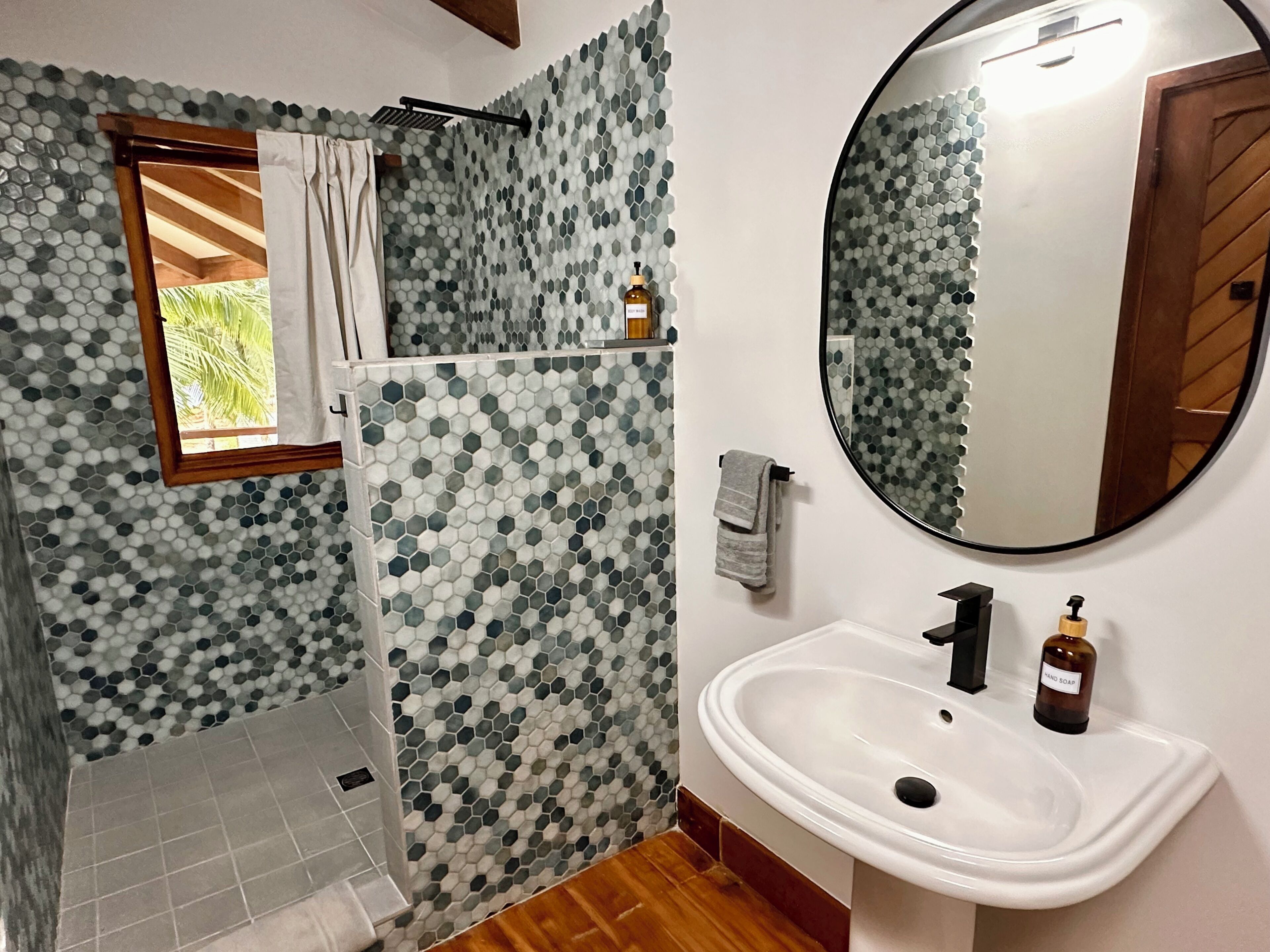 Un baño con ducha, lavamanos, espejo y ventana con vistas a palmeras.