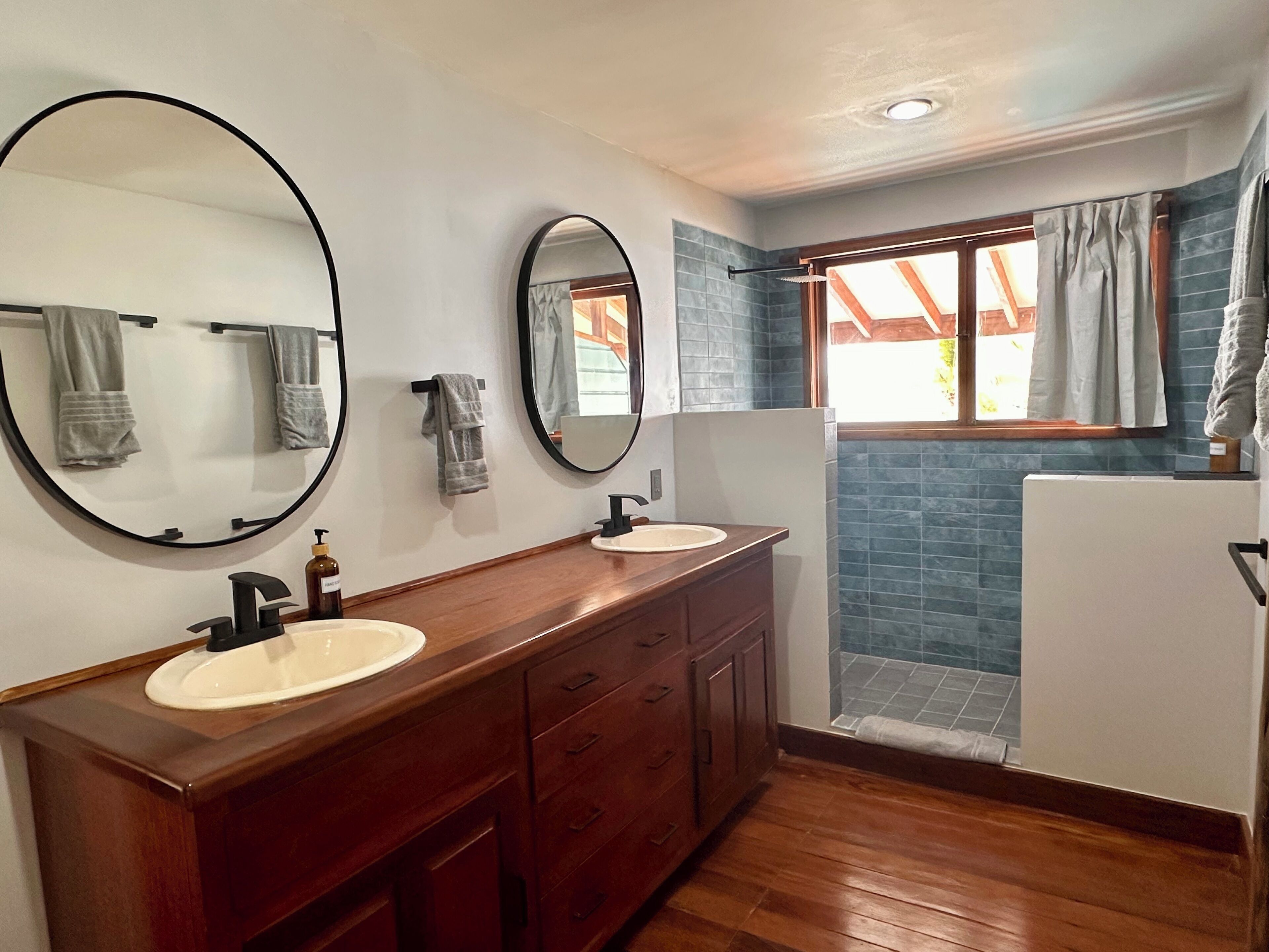 Un baño con dos lavamanos, tocador de madera, espejos grandes y una zona de ducha con azulejos azules.