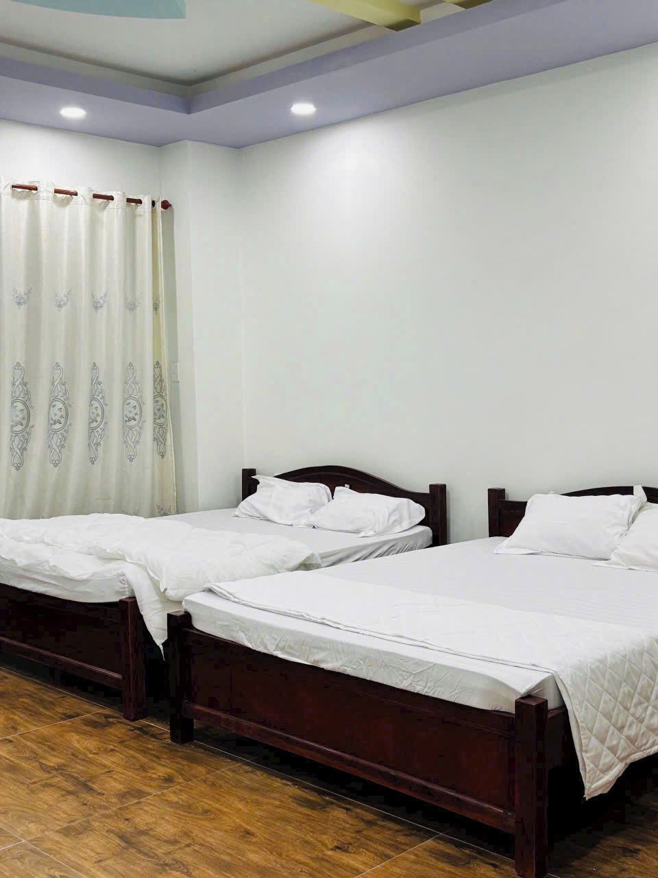 Habitación de hotel con dos camas de madera, ropa de cama blanca, suelo de madera y una cortina blanca con estampado floral.