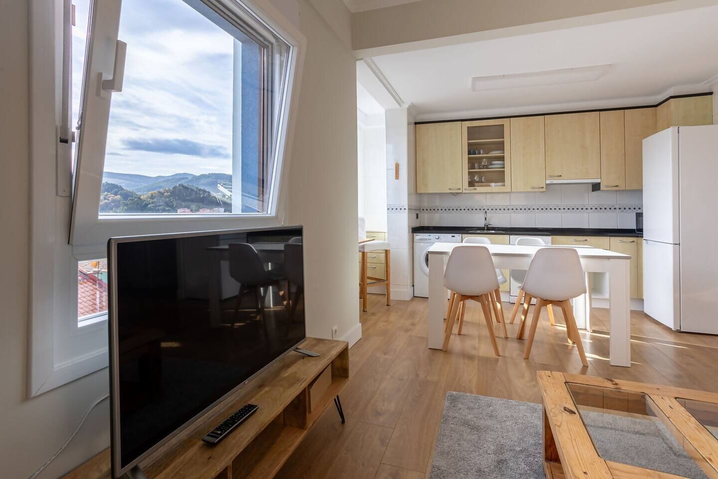 Moderni keittiö, jossa on puiset kaapit, valkoiset tuolit ruokailutilassa ja televisio puisen jalustan päällä.