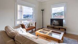 Una sala de estar con un sofá, una mesa de centro, un televisor y vistas a un paisaje urbano a través de grandes ventanales.