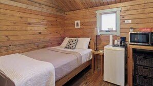 Een houten kamer met een bed, een tafeltje met een lamp, een magnetron en een koelkast.
