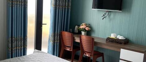 A hotel room with a bed, a desk with two chairs, a television mounted on the wall, and a window with curtains.